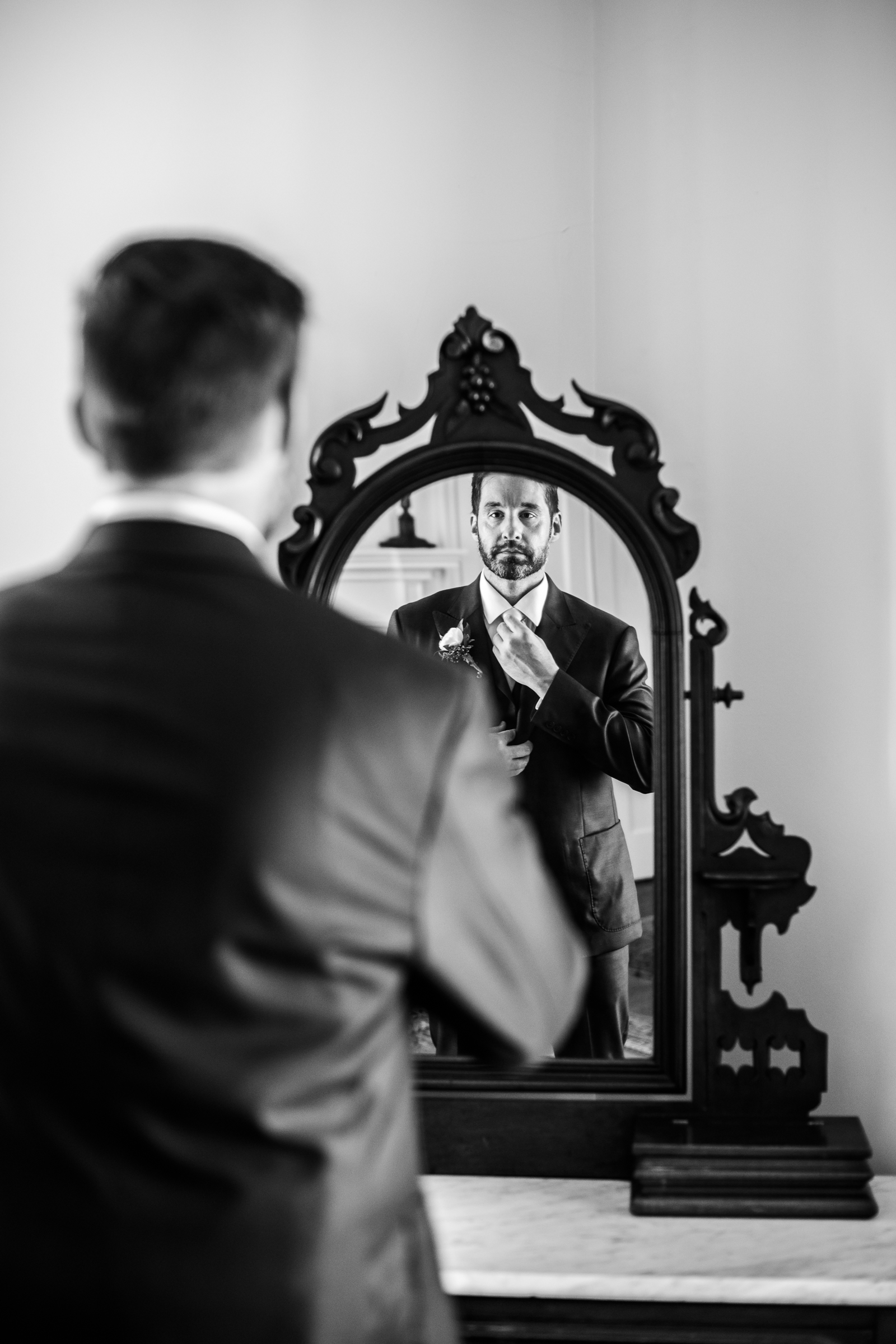 A classic black and white portrait of a groom straightening his tie in front of a mirror in Loudoun County Virginia