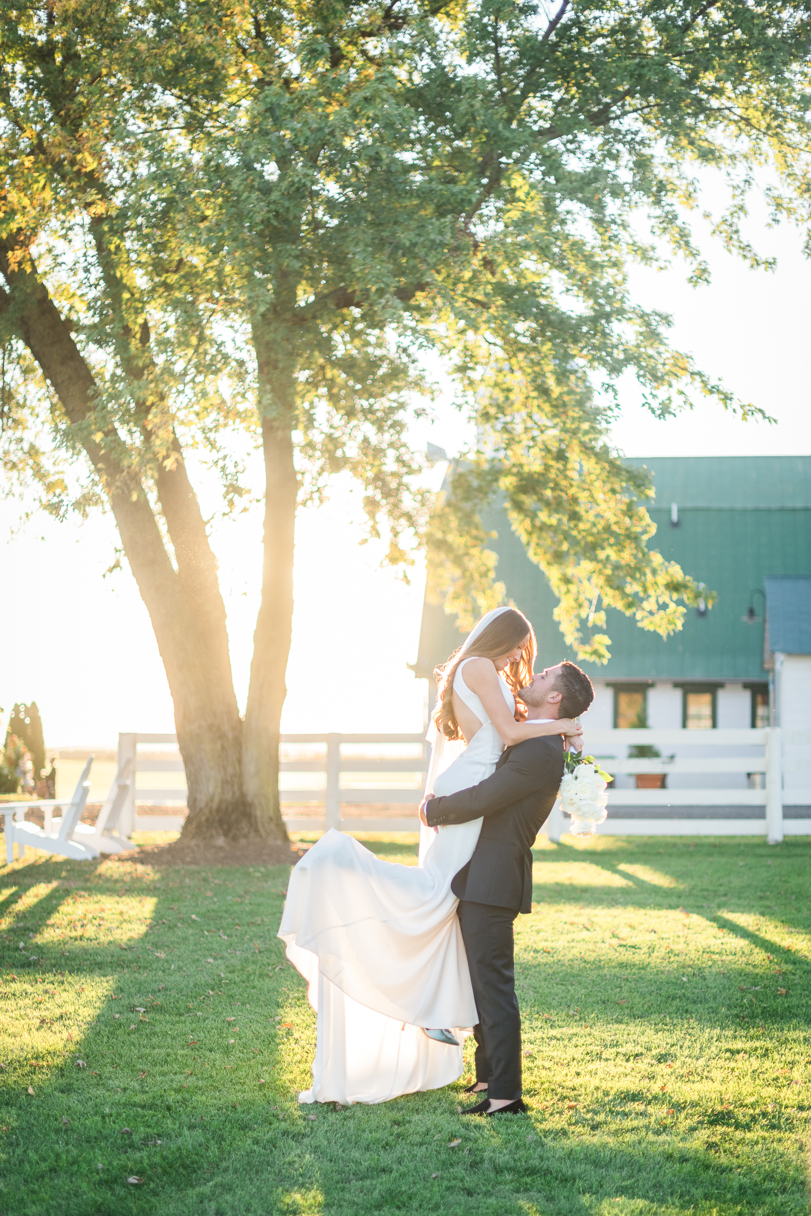 Bride lifted by groom at sunset near white fence at Bluebird Manor wedding venue