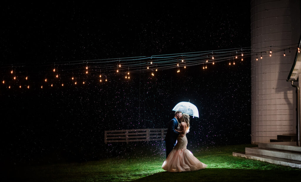 Bride and groom kissing under a clear umbrella at Bluebird Manor during a rainy night wedding.